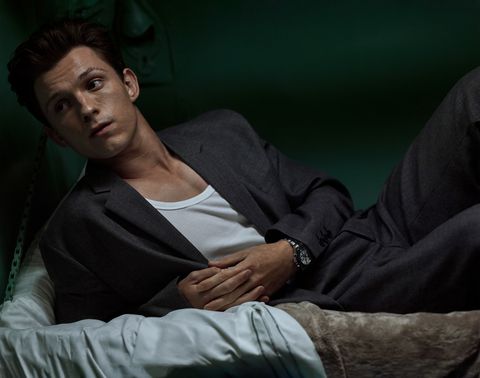 Tom Holland wears a dark-gray suit, white singlet, and lies down on a bed for an editorial shoot