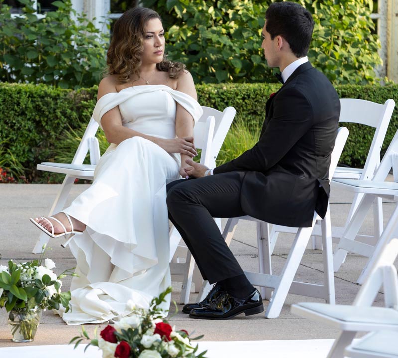 Mallory and Salvador are sitting down on chairs on their wedding day. She is wearing a white dress and he is wearing a black suit. They are both hispanic. 